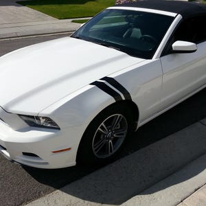 Puede incluir: Ford Mustang convertible blanco con rayas de carreras negras en los guardabarros delanteros. El coche tiene una capota blanda negra y ruedas negras.