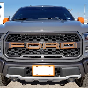 以下が含まれることがあります： オレンジのグリルとオレンジのアクセントが付いたグレーのフォードF-150ピックアップトラック。トラックはハニカムパターンで黒いグリルとオレンジの文字で「FORD」と書かれています。トラックはシルバーの牽引フック付きの黒いバンパーとオレンジの縁取りのある黒いナンバープレートフレームを持っています。