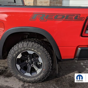 May include: A red pickup truck with a black "Rebel" decal on the side. The truck has black fender flares and a black rear bumper. The truck is sitting on a black asphalt surface. The truck has a black tire with a Goodyear Wrangler tire.