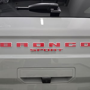 Puede incluir: Un port&oacute;n trasero de veh&iacute;culo de color gris claro con las letras rojas BRONCO SPORT centradas horizontalmente. Un limpiaparabrisas trasero negro est&aacute; colocado en la parte superior y una manija rectangular con c&aacute;mara integrada se encuentra debajo.