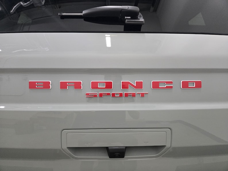 Puede incluir: Un port&oacute;n trasero de veh&iacute;culo de color gris claro con las letras rojas BRONCO SPORT centradas horizontalmente. Un limpiaparabrisas trasero negro est&aacute; colocado en la parte superior y una manija rectangular con c&aacute;mara integrada se encuentra debajo.