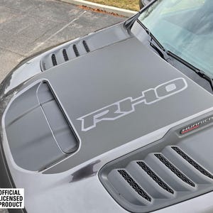 Pode incluir: Um capô de carro preto com um decalque "RHO" preto e prateado e um decalque "HURRICANE" preto. O capô tem uma grande ventilação e várias ventilações menores.
