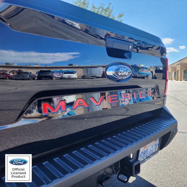 May include: Black Ford Maverick truck tailgate with red "MAVERICK" lettering and a blue Ford emblem. The truck's bumper and hitch are visible, with a "Ford Official Licensed Product" sticker in the corner.