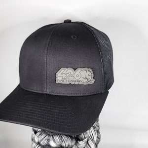 May include: Black trucker hat with a curved brim and mesh back. The hat features a silver emblem with the text "426 MOPAR" and a stylized design. The hat is displayed against a white background.