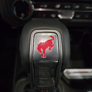 Op de afbeelding: Een zwarte versnellingspookknop voor auto's met een metallic zilveren inleg in het midden en een felrood embleem van een steigerend paard. De versnellingspook heeft een gladde, matzwarte afwerking aan de zijkanten en een glanzend oppervlak.