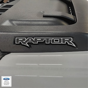 May include: A close-up of a black truck hood with a silver "RAPTOR" emblem. The emblem is raised and has a black outline. The hood is textured and has a slight sheen.
