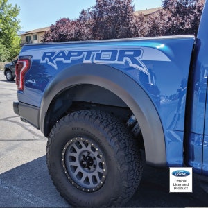 May include: A blue Ford Raptor pickup truck with a black fender and a gray wheel with black accents. The truck has a "RAPTOR" decal on the side and a Ford logo with the text "Official Licensed Product".