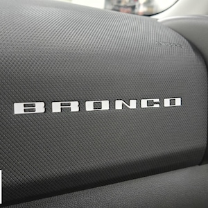 May include: Close-up of a car dashboard with the word "BRONCO" in silver block letters. The dashboard is dark gray with a textured pattern. A small Ford logo is in the lower left corner.