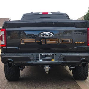 May include: Black Ford F-150 Tremor truck with orange lettering. The truck has a Colorado number plate and dual exhaust pipes. The Ford logo is visible above the F-150 lettering. The truck is parked on a paved surface.