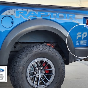 Pode incluir: Uma caminhonete Ford Raptor azul com um adesivo "RAPTOR" preto e cinza no lado. A caminhonete tem um adesivo "FP" preto e cinza com uma bandeira americana no para-lama traseiro. A caminhonete tem um pneu preto com uma roda cinza.