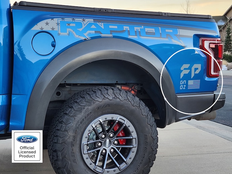 Pode incluir: Uma caminhonete Ford Raptor azul com um adesivo "RAPTOR" preto e cinza no lado. A caminhonete tem um adesivo "FP" preto e cinza com uma bandeira americana no para-lama traseiro. A caminhonete tem um pneu preto com uma roda cinza.
