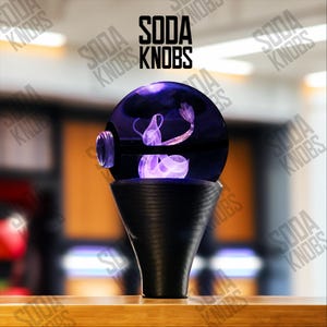 Pode incluir: Um botão decorativo para torneira de refrigerante com um design de água-viva roxa dentro de um vidro redondo e transparente. O botão está sobre uma base preta em forma de cone. O texto "SODA KNOBS" é exibido acima.