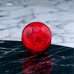 Pode incluir: Um peso de papel de vidro vermelho com uma rosa vermelha dentro. O peso de papel está sobre uma superfície de mármore preto.