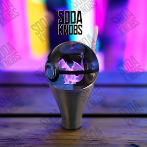 Puede incluir: Un pomo de cambio personalizado con una parte superior esférica transparente que presenta un diseño de dragón púrpura. El pomo tiene una base plateada y el texto "SODA KNOBS" es visible en el fondo. El diseño está iluminado con luz púrpura.