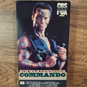May include: A VHS tape of the film Commando starring Arnold Schwarzenegger. The cover features a portrait of the actor in a black tactical vest holding a grenade, with the text CBS FOX VIDEO at the top and SCHWARZENEGGER COMMANDO at the bottom.
