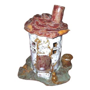 May include: A ceramic house sculpture featuring a white cylindrical body with cut-out windows, a textured reddish-brown roof with a chimney, and a base decorated with small brown figures and a stump, all finished in a glossy, multi-coloured glaze.
