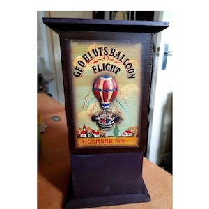 May include: A wooden box with a painted image of a hot air balloon with the text "GEO BLUTS BALLOON FLIGHT RICHMOND 1816". The box is dark purple with a rectangular shape and a flat top.