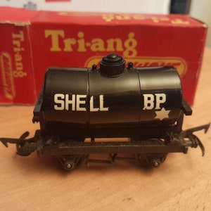 May include: A black vintage Tri-ang Shell BP tanker train car with white lettering and a star graphic. The toy train car is on a wooden surface with a red box in the background.