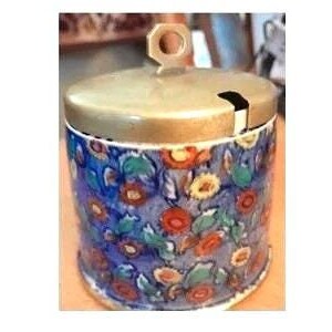 Peut inclure: Petit pot cylindrique en céramique avec couvercle et poignée en laiton. Le pot est décoré d'un motif floral dans les tons de bleu, rouge, orange et vert. Le couvercle a une petite ouverture pour une cuillère.