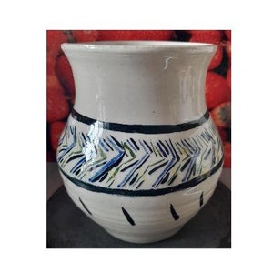 Puede incluir: Un jarrón de cerámica blanco con una abertura ancha y un cuerpo redondeado. El jarrón presenta una banda de patrones en zigzag azules, verdes y amarillos entre dos bandas azul oscuro. Líneas negras verticales decoran la parte inferior.