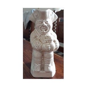 Puede incluir: Figura de cerámica color crema de un chef sosteniendo una bolsa. El chef lleva un sombrero alto y un uniforme detallado. La figura tiene un diseño caprichoso y caricaturesco y se apoya sobre una base rectangular. El fondo está borroso, con muebles de madera visibles.