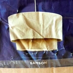 May include: A handmade, two-tiered lampshade crafted from light brown paper, suspended by a thin white rod. The lampshade has a layered design, with the top tier slightly smaller than the bottom. The background is a dark blue, textured surface.