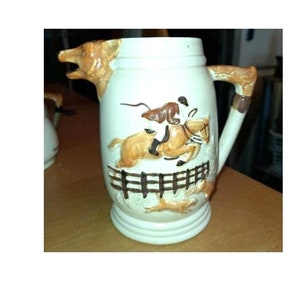 May include: Cream ceramic pitcher with a hunting scene. The pitcher has a brown handle and a spout shaped like a dog's head. The design depicts a horse and rider jumping a fence, with a fox and dogs in pursuit.