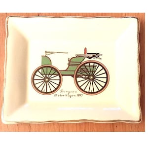 May include: Rectangular decorative dish with a cream-colored background and a gold-colored rim. The dish features a green and black illustration of a vintage Duryea Motor Wagon from 1895. The text "Duryea's Motor Wagon 1895" is below the illustration.