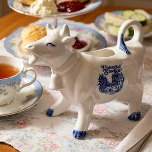 Peut inclure: Un pot à lait en céramique blanche en forme de vache avec des détails peints en bleu sur les sabots, la queue et les yeux. Le côté présente un motif bleu d'un château et le texte Universal Studios. Le pot possède une cloche et une anse.