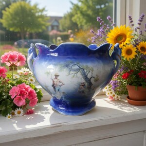 May include: A blue and white ceramic bowl with a scalloped rim and two side handles. The exterior features a painted scene of a person standing near a tree. The bowl has a glossy finish and a wide, rounded body that tapers towards the base.