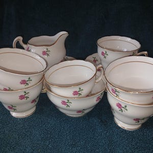 May include: A collection of white porcelain teacups and a creamer pitcher, each decorated with small pink rose motifs and green leaves. The pieces feature scalloped edges and delicate gold trim along the rims and handles, creating a cohesive set.