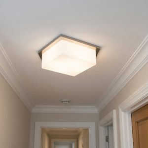 May include: A square flush mount ceiling light fitting featuring a metallic frame and a frosted white glass shade that emits a soft, diffused glow. The fitting is mounted directly to a white ceiling in a hallway with crown moulding.