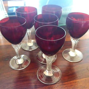 May include: A set of five wine glasses featuring deep red bowls and clear, twisted glass stems. Each glass has a rounded bowl shape and a circular base, showcasing a consistent design across the collection with transparent stems and dark red glass.