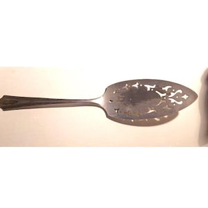 Peut inclure: Une pelle à gâteau en métal argenté dotée d'un long manche et d'une large lame en forme de pique. La lame est ornée de motifs découpés symétriques et complexes sur ses bords, tandis que le manche présente un design simple et effilé.