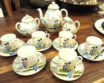 May include: A vintage porcelain tea set with a Japanese geisha design. The set includes a teapot, sugar bowl, creamer, and six teacups with saucers. The design features geishas in colorful kimonos, set against a landscape with mountains and trees.