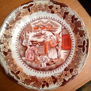 May include: A decorative plate with a brown and white floral border and a central scene depicting figures in traditional clothing. The scene includes buildings, a boat, and a bird. The plate has a scalloped edge.