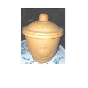 May include: A tan ceramic garlic keeper with a lid. The keeper has a bulbous shape and a carved garlic design on the front. The lid has a small knob. The keeper is on a plate with a floral pattern.