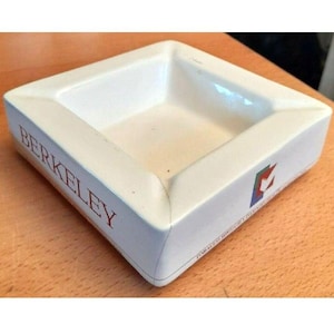 May include: White ceramic ashtray with a square shape and angled sides. The word "BERKELEY" is printed in red on one side. A logo with red, green, and blue colors is on another side. The ashtray is on a light brown surface.