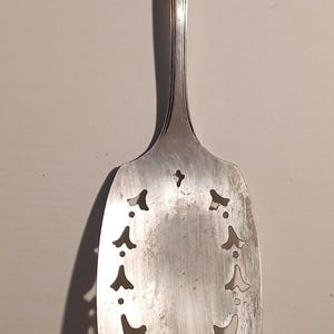 Puede incluir: Cuchara de servir plateada con un diseño decorativo calado en la superficie de servir. El mango tiene ranuras verticales y un pequeño elemento decorativo en la parte superior. Un utensilio de cocina clásico.