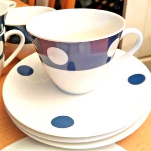 May include: White teacup and saucer set with a navy blue band and large white polka dots. The saucer has three navy blue polka dots. The teacup has a white handle. The set is on a wooden surface.