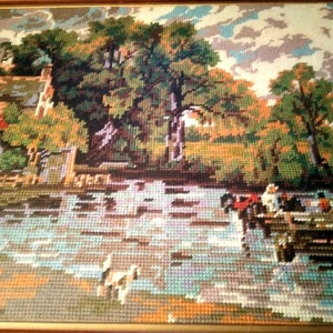 May include: A framed needlepoint landscape showing a river scene with a bridge, buildings, and trees. The artwork uses greens, browns, and blues, creating a detailed and textured image. The frame is a warm brown colour.