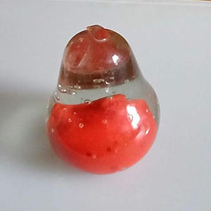 May include: A clear glass paperweight with a red, pear-shaped interior. The glass has small air bubbles throughout, and the red portion is textured. The paperweight is on a white surface.