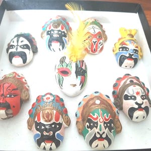 Peut inclure: Collection de neuf petits masques d'opéra chinois décoratifs dans une boîte noire. Les masques sont peints à la main avec des couleurs vives, présentant des motifs complexes et des expressions faciales. Une plume jaune est placée au centre.