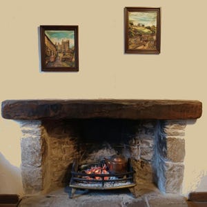 May include: A stone fireplace with a thick, dark brown wooden mantelpiece. Inside the hearth, a metal grate holds glowing embers and a copper kettle. Two framed landscape paintings hang on the light-colored wall above the fireplace mantel.