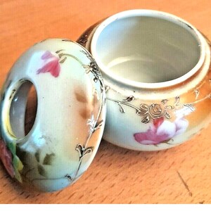 May include: An open, antique porcelain trinket box with a lid. The box and lid are decorated with pink roses and gold accents. The lid has a handle with a hole. The box is on a wooden surface.