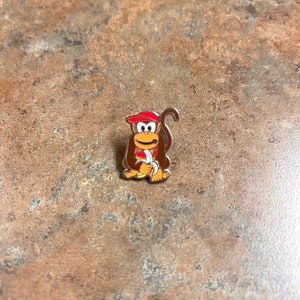 May include: A metal pin featuring a cartoon monkey wearing a red hat and holding a banana. The monkey is brown with a white face and a yellow banana.