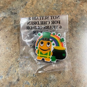 Pode incluir: Um personagem de desenho animado amarelo usando um chapéu verde e segurando um pote de ouro. O personagem está em pé na frente de um arco-íris com trevos de quatro folhas. O texto "Não adequado para crianças menores de 6 anos" está impresso na embalagem.