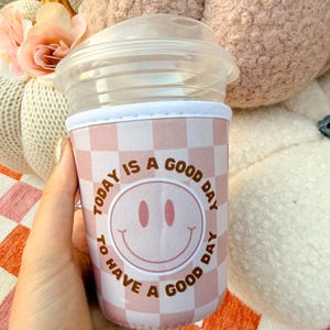 Hoy es un buen día para tener un buen día. Funda de café con cara sonriente, cara feliz y diseño a cuadros beige.