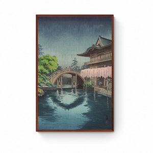 May include: A framed print depicting a Japanese landscape with a wooden arched bridge over water. A traditional building with a curved roof and hanging flowers is on the right. The color scheme includes blues, greens, and browns.