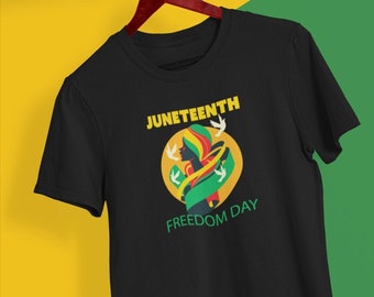 Può includere: Maglietta nera con un design grafico di una donna con una fascia gialla e verde, circondata da colombe bianche. Il testo "Juneteenth Freedom Day" è stampato sulla maglietta.